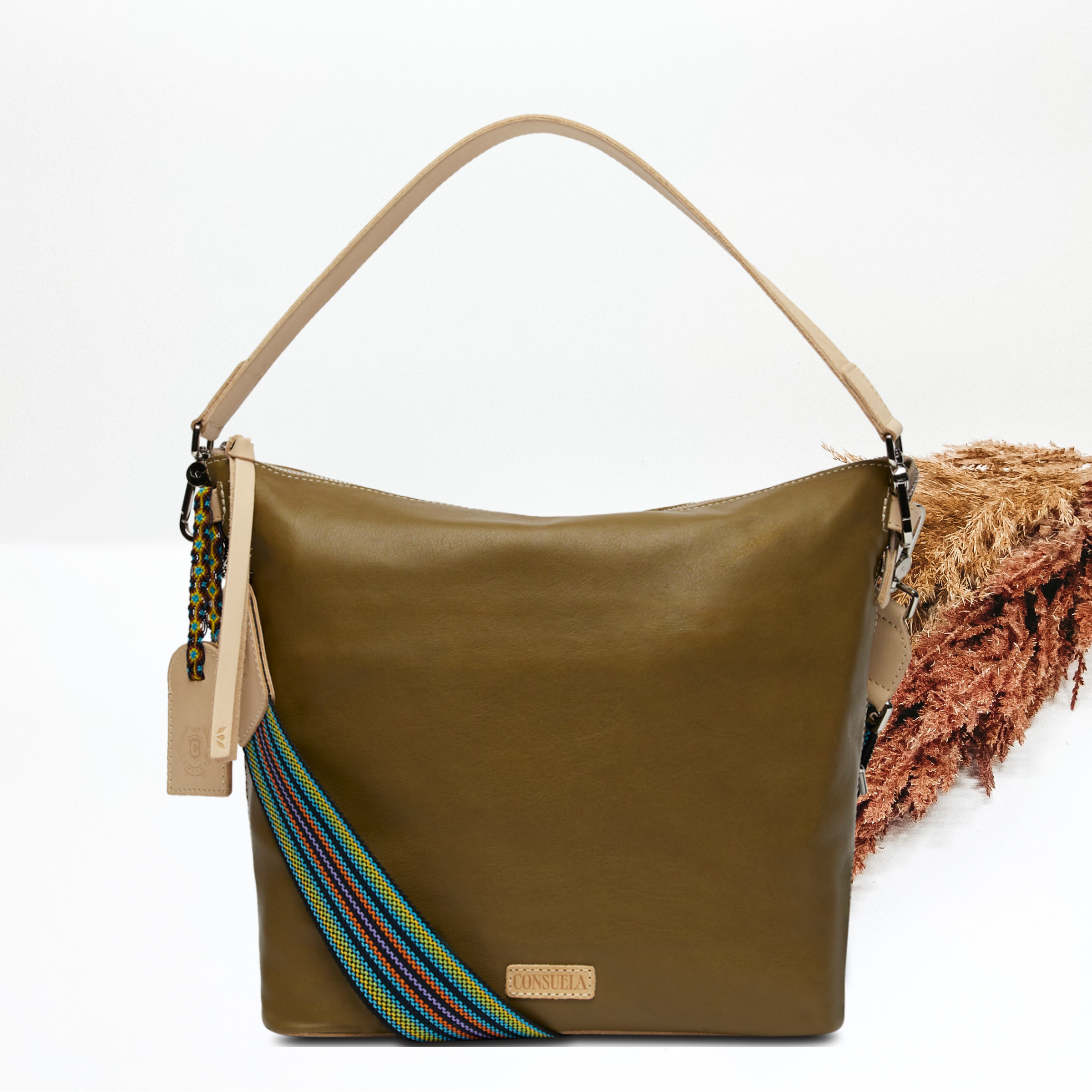 Pictured on a white background is olive green leather hobo bag with a light tan shoulder strap. This bag also includes a striped crossbody strap., light tan tassel on the zipper and light tan charm. 
