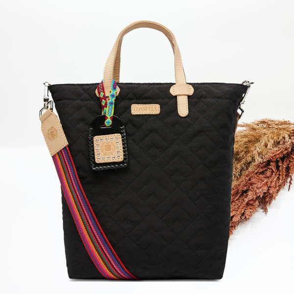Pictured on a white background is a black essential tote bag that has a diamond stitched print. This purse includes a black and tan charm, a striped purse strap, and light tan handles. 
