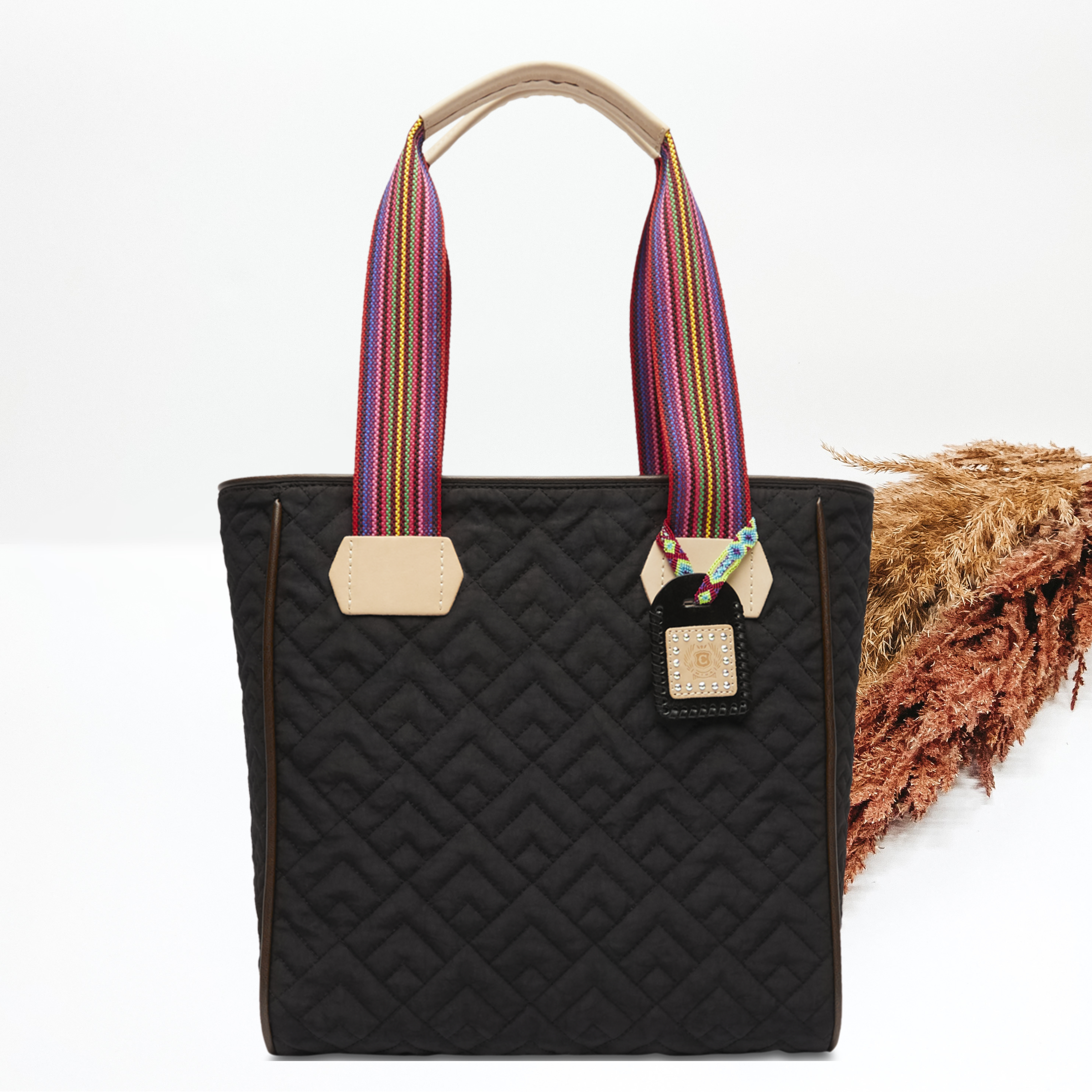 Pictured on a white background is a black classicbag that has a diamond stitched print. This purse includes a black and tan charm and striped straps with leather handle holds. 