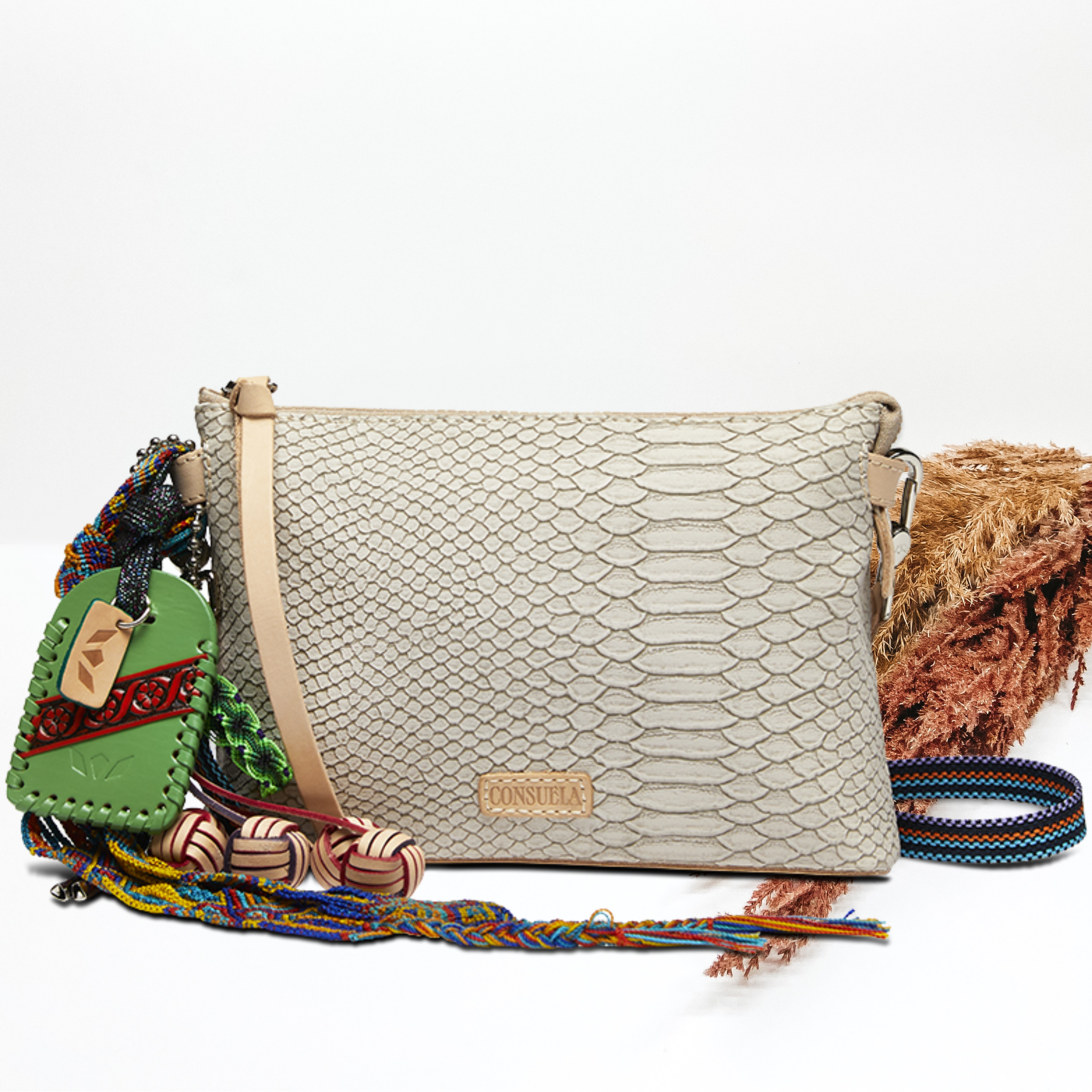 Pictured on a white background is a white snake print midtown crossbody purse. This purse includes multiple, multicolored charms and a striped strap. 