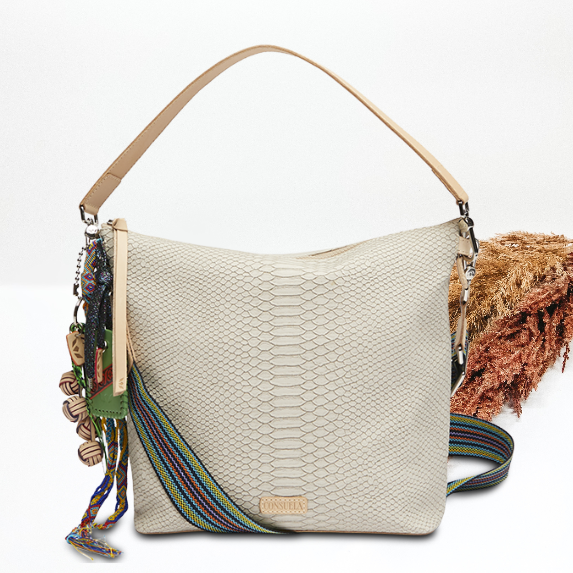 Pictured on a white background is a white snake print hobo bag. This purse includes multiple, multicolored charms and a light tan shoulder strap 