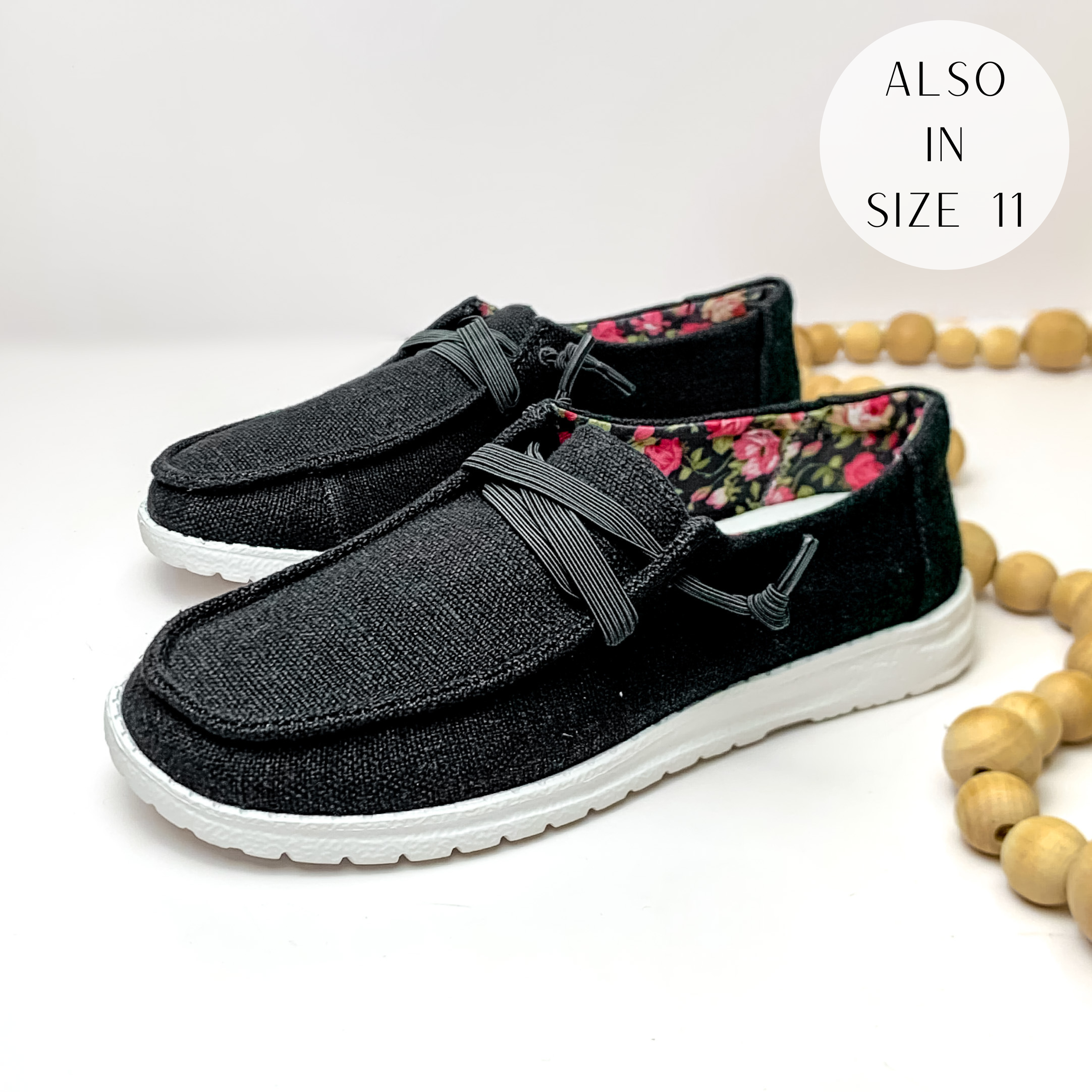 Pictured are a pair of black canvas shoes with a white sole, black laces, and a pink rose pattern inside the shoe. These shoes are pictured on a white background with tan beads on the right side.