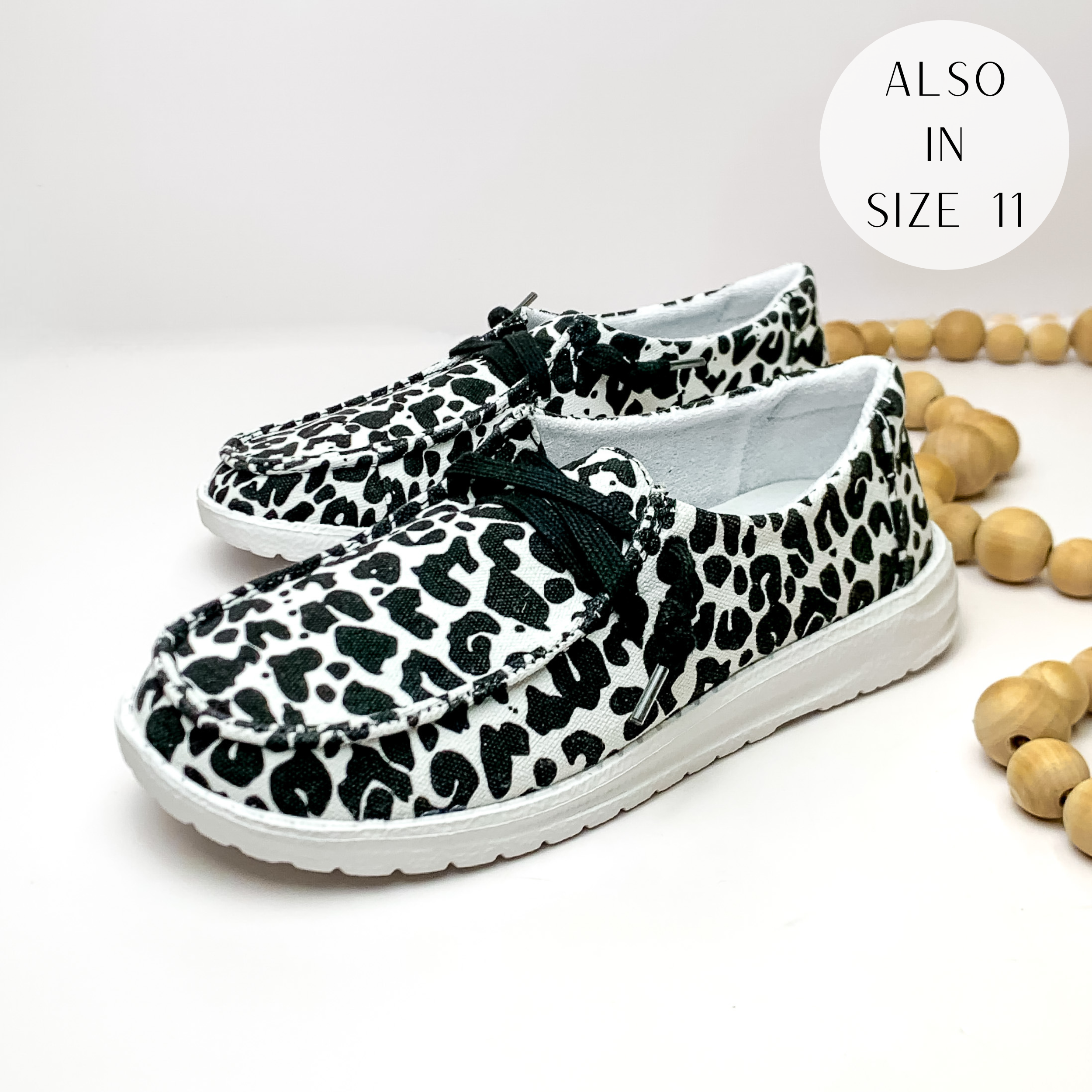 White, slip on loafers with a black leopard print and black laces. These shoes are pictured on a white background with tan beads on the right hand side. 