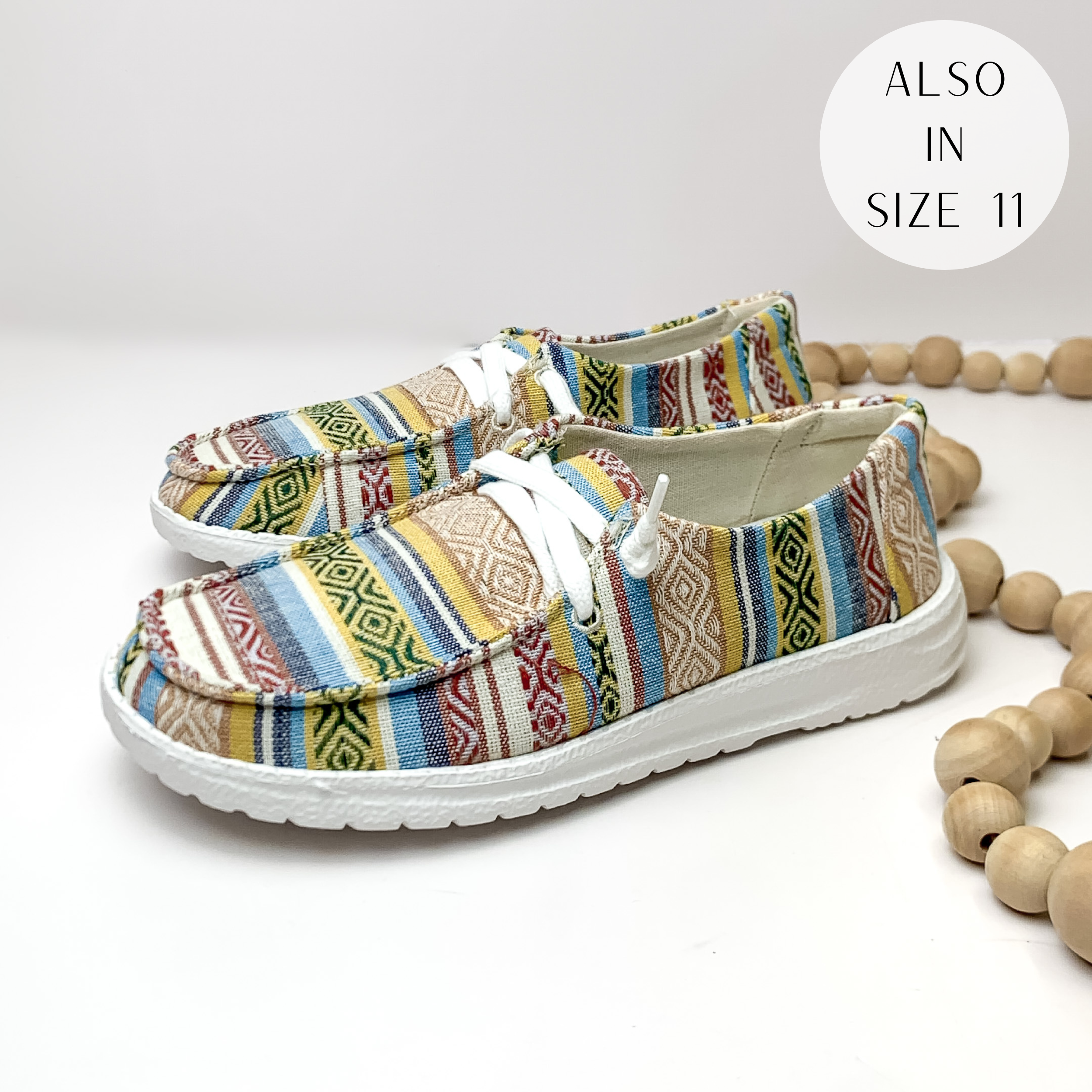 Pictured are a pair of cream canvas shoes with a multicolored tribal print with a white sole and white laces. These shoes are pictured on a white background with tan beads on the right side.