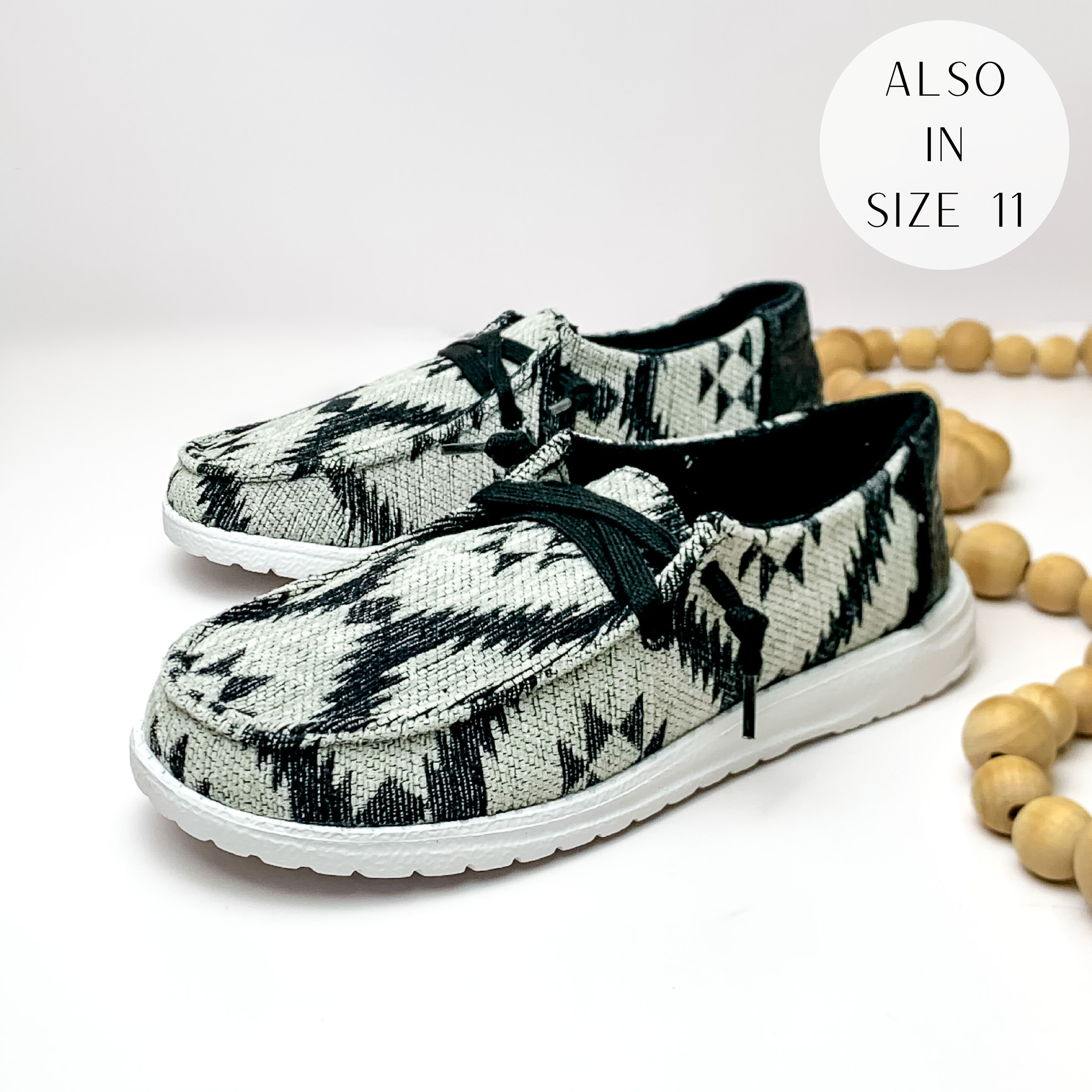 Pictured is a pair of grey canvas shoes with a black aztec print with a white sole and black laces. These shoes also include black, leather tooled heels. These shoes are pictured on a white background with tan beads on the right side.