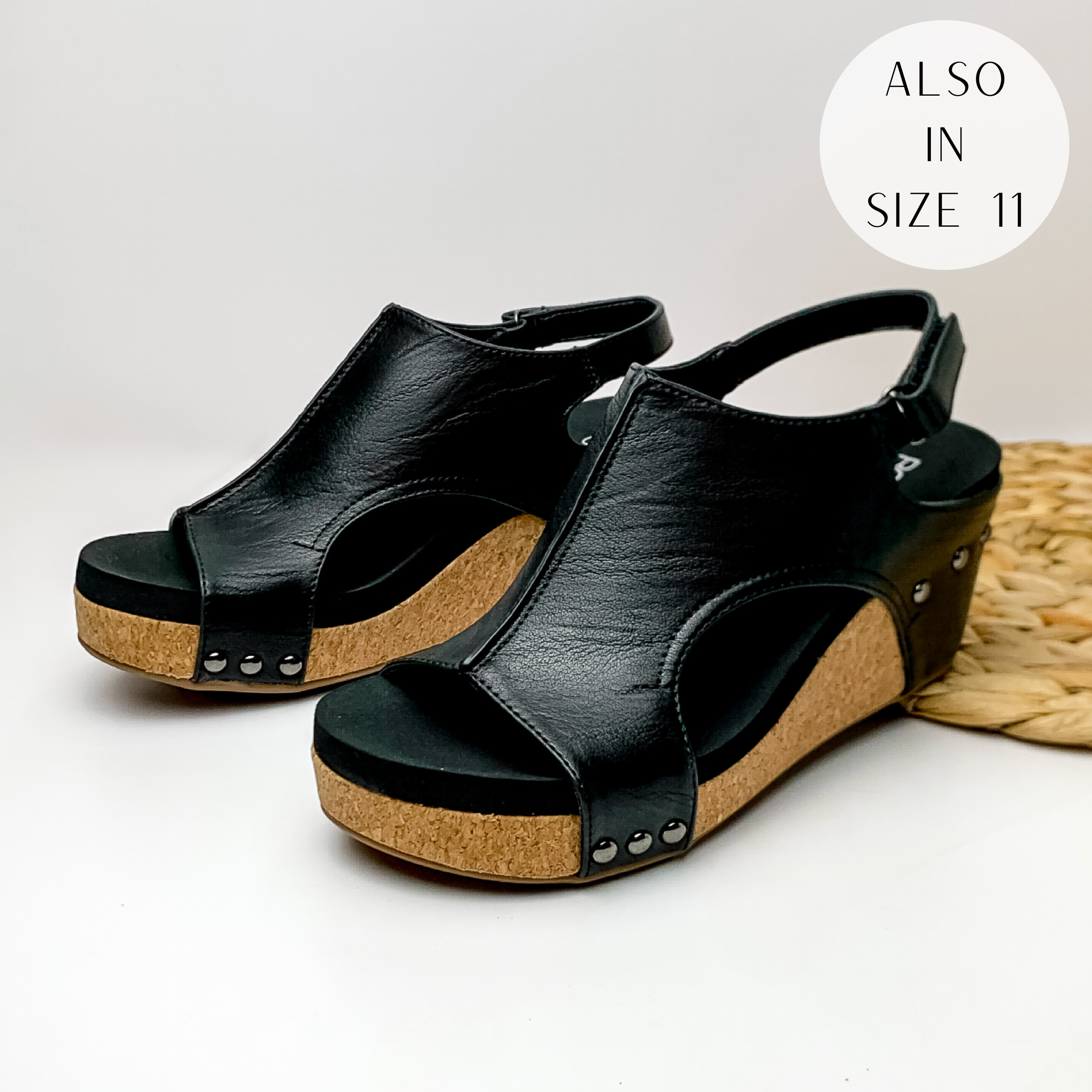 Cork wedges with a black color upper with bronze studs connecting the upper to the wedge. These wedges also include a velco strap. These wedges are pictured on a white background with a basket weave material on the right side of the picture.