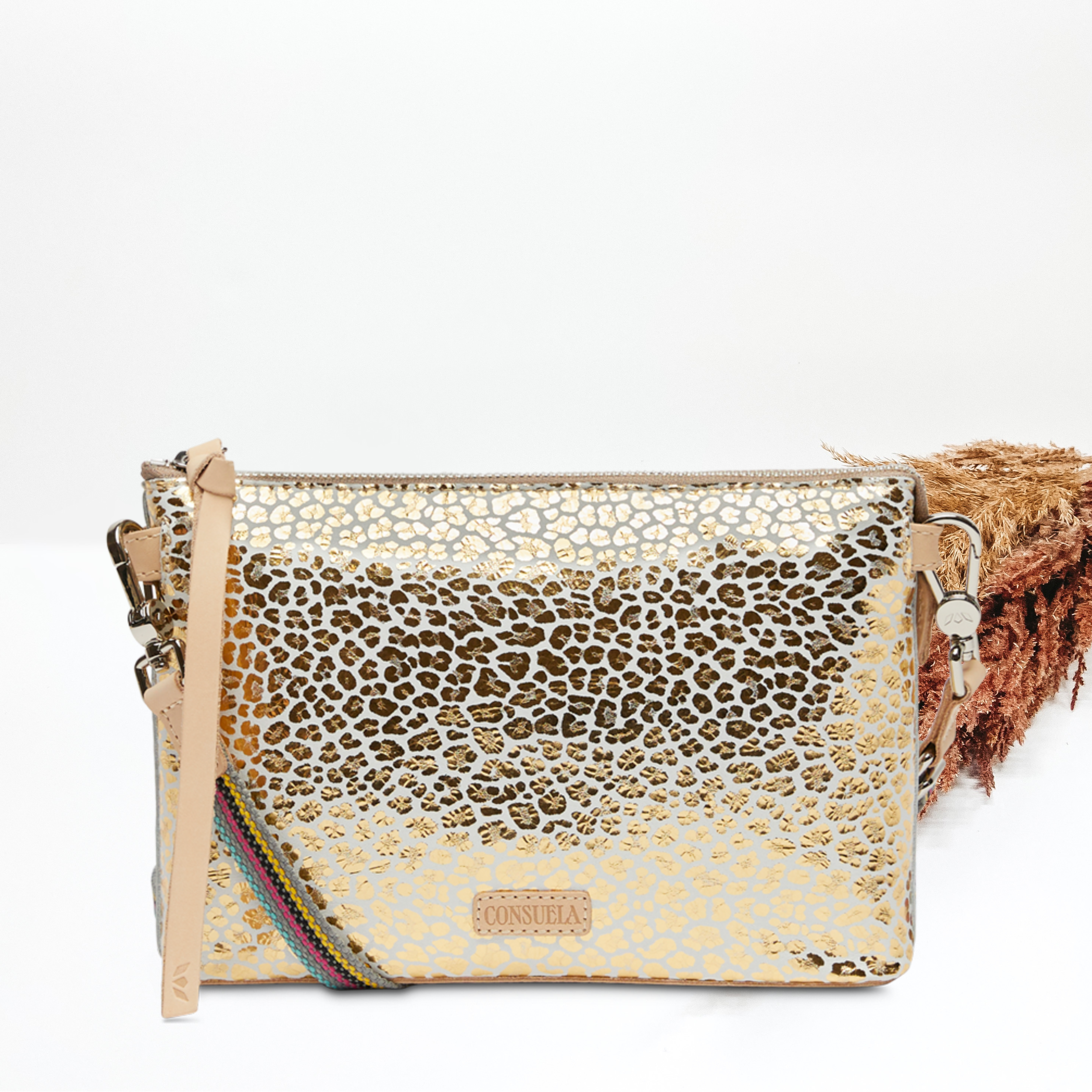 Pictured on a white background is a midtown corssbody purse with a light tan leather zipper pull on the top and a woven strap. This purse has a white with a metallic gold leopard print design. 
