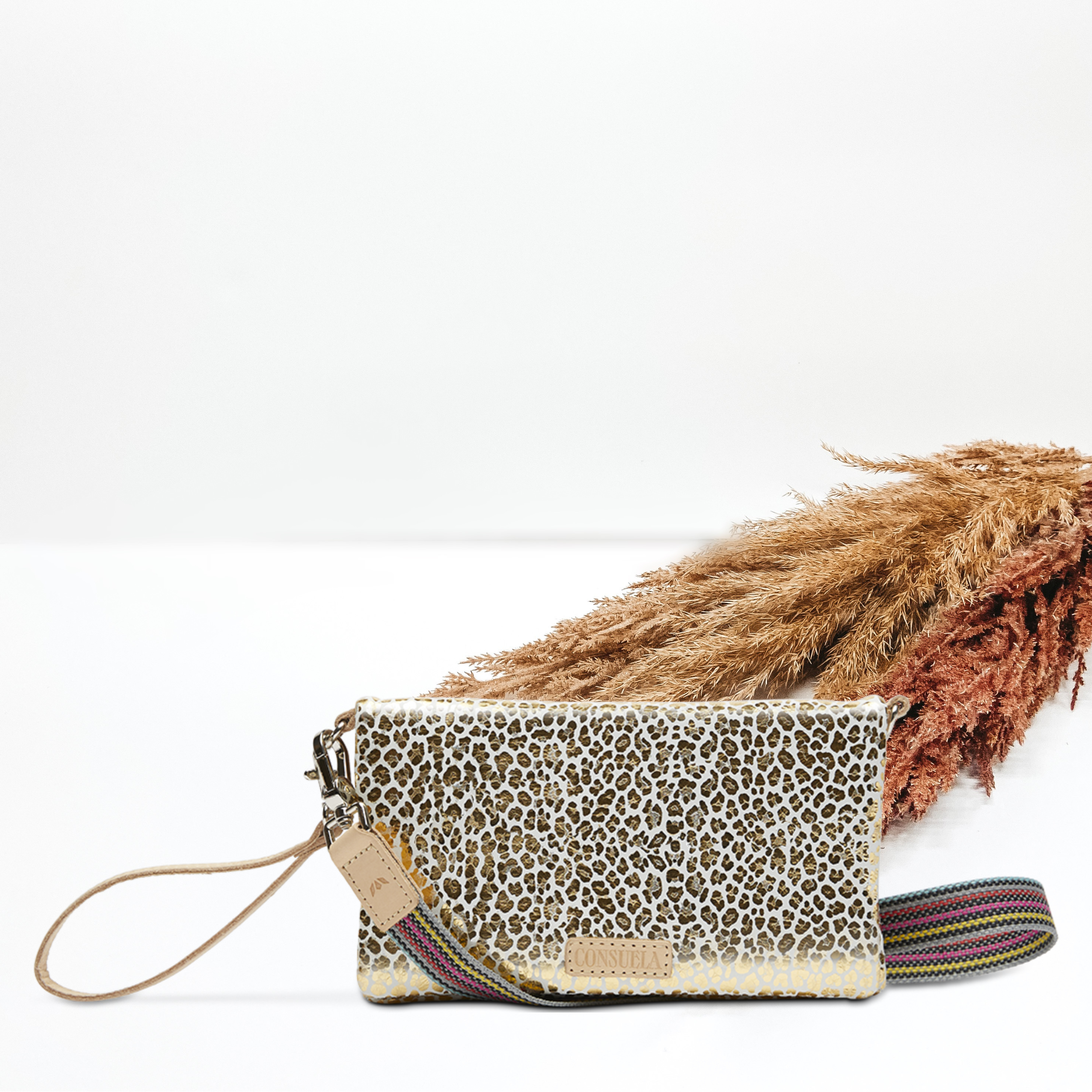 Pictured on a white background is an uptown corssbody purse with a light tan leather wristlet strap and a woven crossbody strap. This purse has a white with a metallic gold leopard print design. 