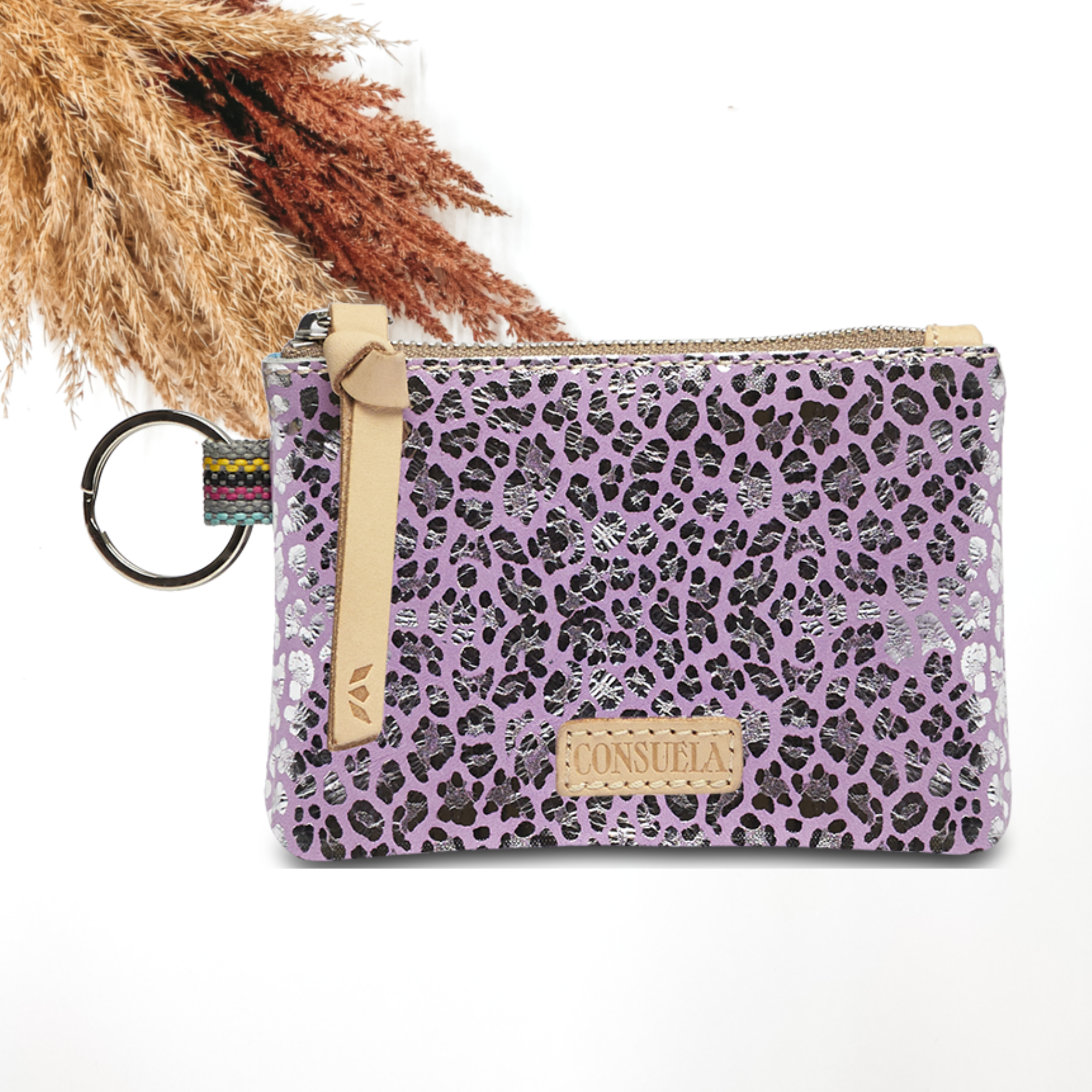 Pictured on a white background is a pouch with a light tan leather zipper pull on the yop and a silver key ring on the side. This pouch has a purple with a metallic silver leopard print design.