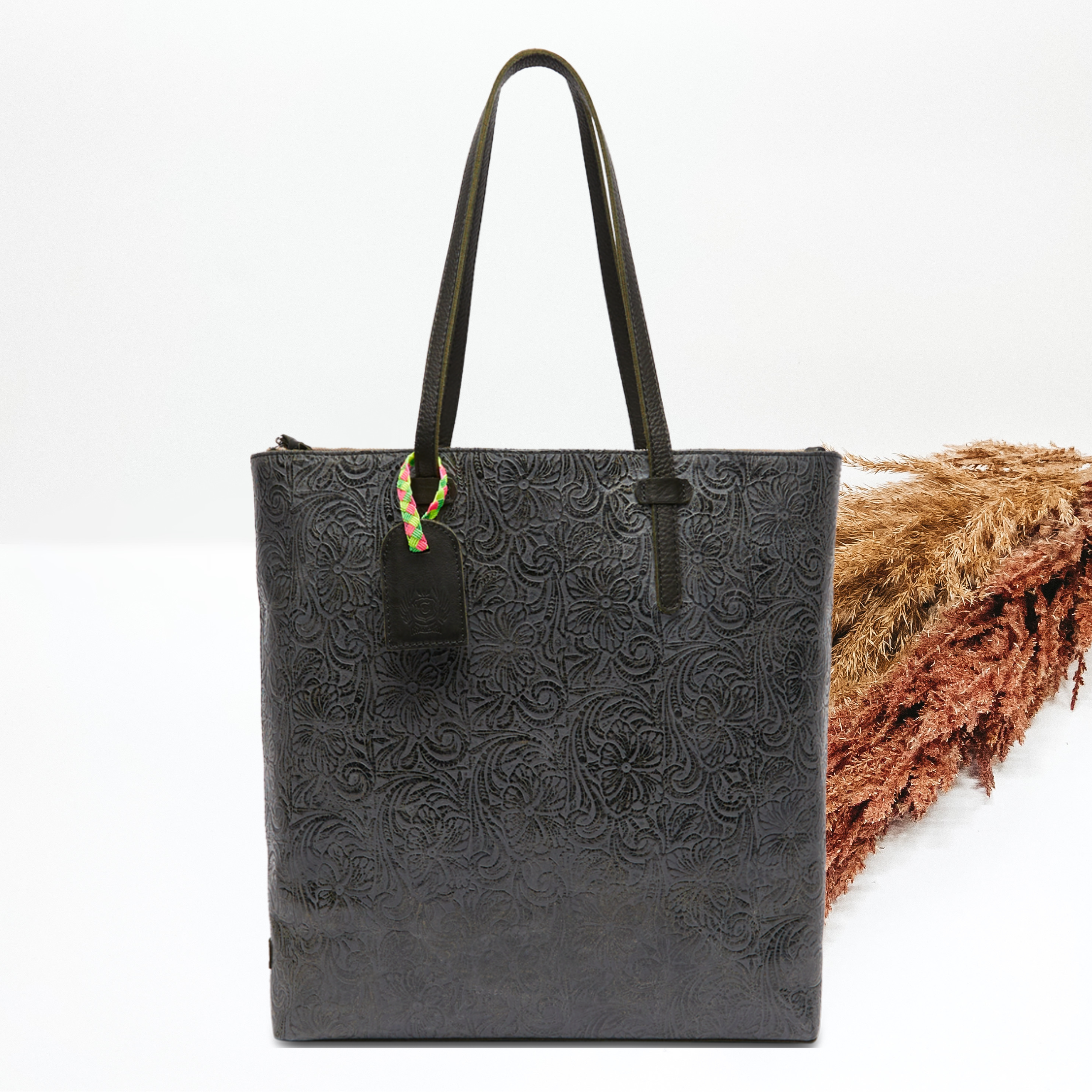 Pictured on a white background is a market tote bag with black leather shoulder straps and a a black luggage tag. This purse has a black leather tooled print.