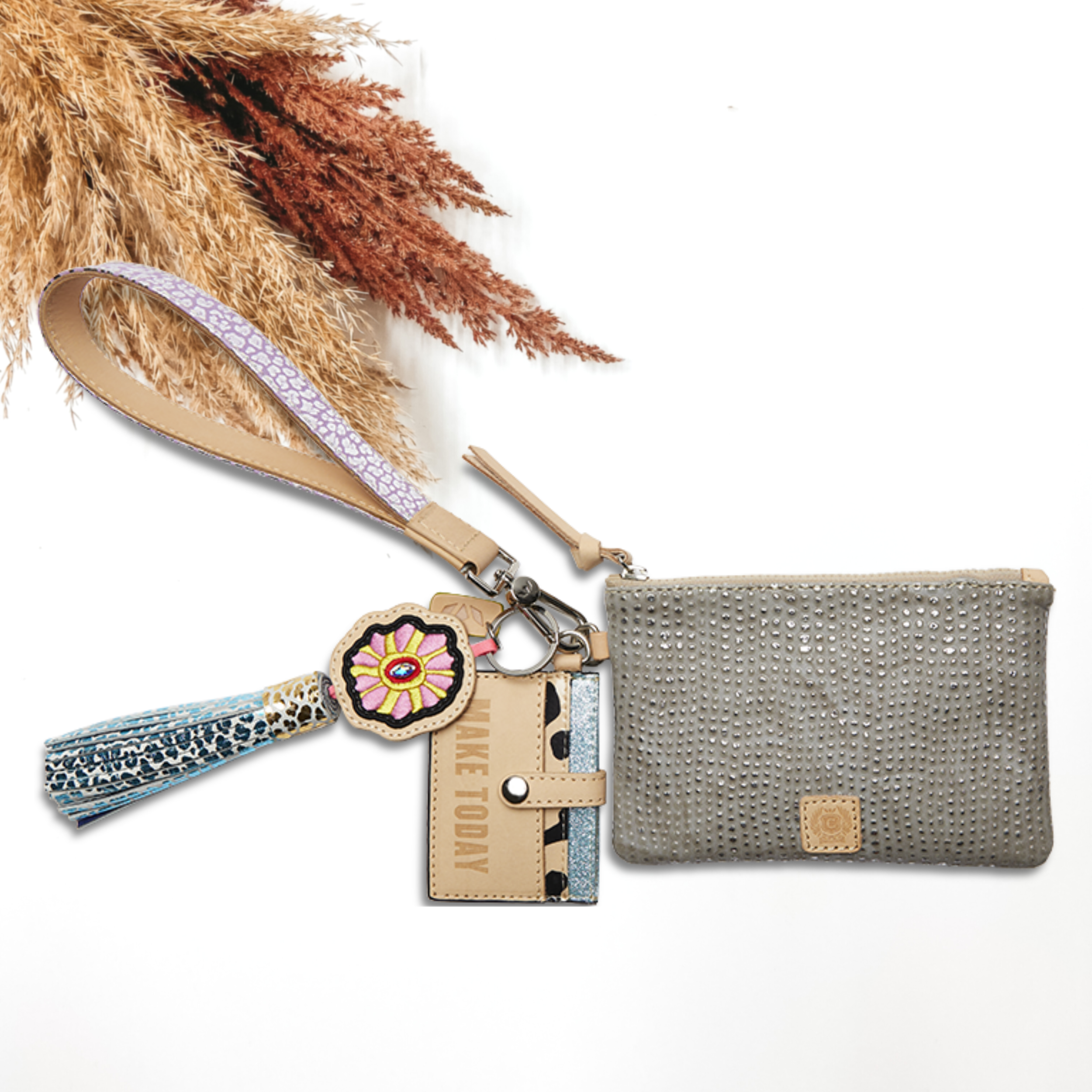 Pictured on a white background is a grey pouch with a light tan and purple wristlet. This pouch also includes a flower charm, light tan tassel on the zipper, and a card holder keychain.
