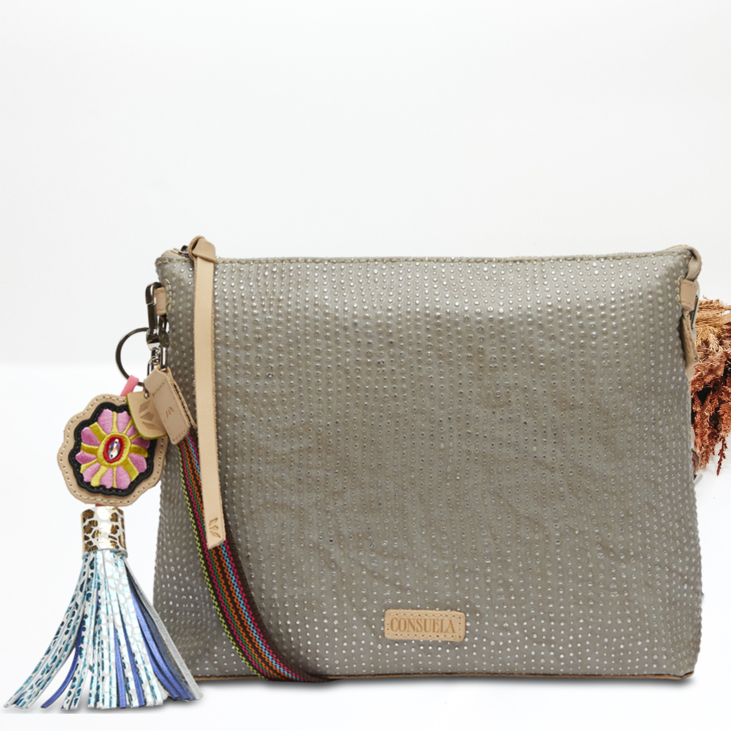 Pictured on a white background is a grey downtown crossbody purse with a silver dotted print. This purse has a woven crossbody strap, a multicolored tassel charm and light tan leather zipper pull.