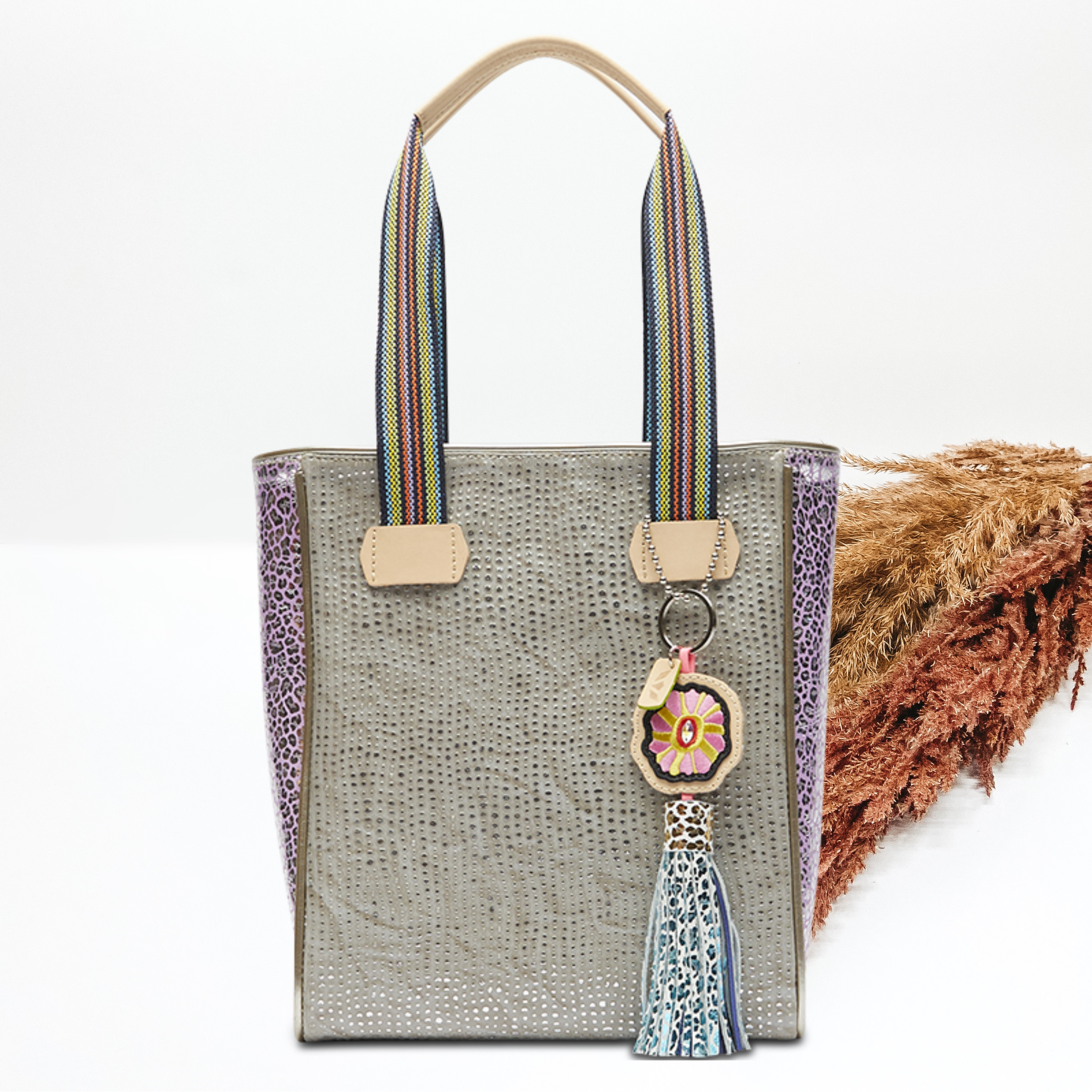 Pictured on a white background is a grey chica tote bag with woven straps and light tan handles. This bag includes a silver dotted print, purple leopard side panels, and a multicolored tassel charm. 