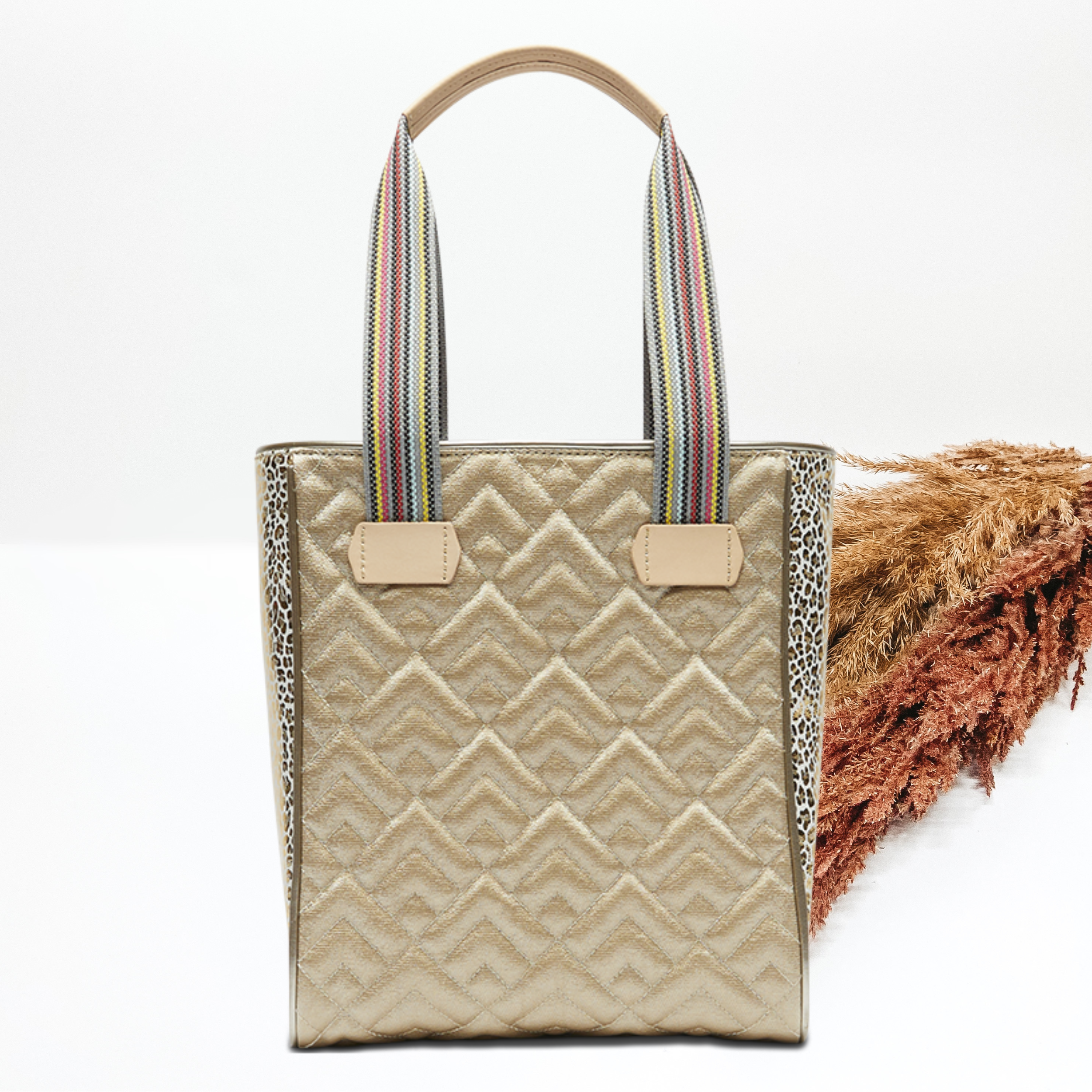 Pictured on a white background is a gold chica tote bag with woven straps and light tan handles. This bag includes a diamond stitched pattern and gold leopard side panels. 