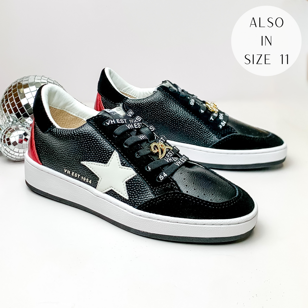 Pictured is a pair of black tennis shoes with black suede, red shimmer, and white detailing. These shoes include a red shimmer heel, a white star on the side, and black laces. These shoes are pictured on a white background with disco balls on the left side