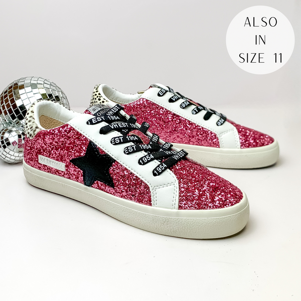 Pictured is a pair of pink glitter tennis shoes with black, cheetah print, and white detailing. These shoes include a white and black cheetah print heel, a black star on the side, and black laces. These shoes are pictured on a white background with disco balls on the left side