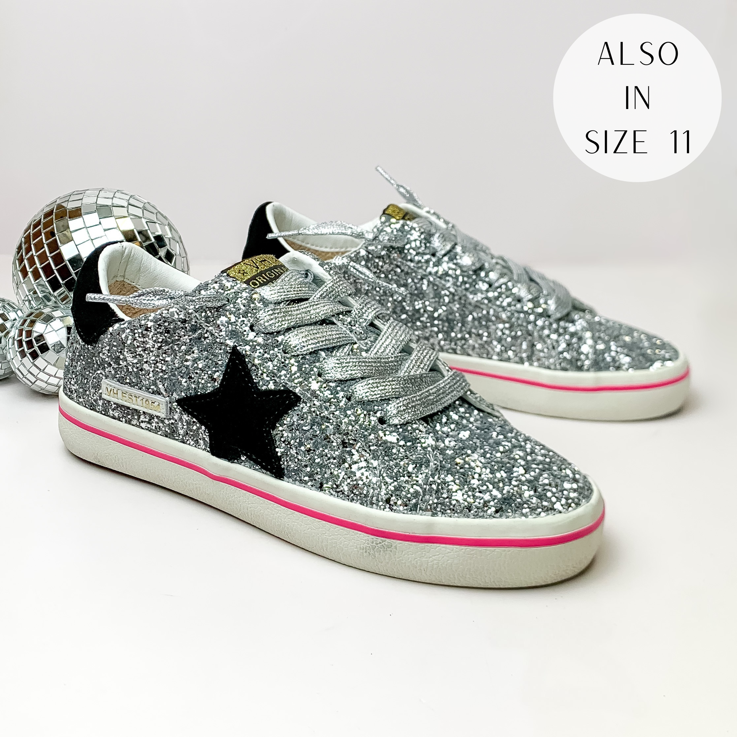 Pictured is a pair of silver glitter tennis shoes with black detailing. These shoes include a black heel, a black star on the side, and silver laces. These shoes are pictured on a white background with disco balls on the left side