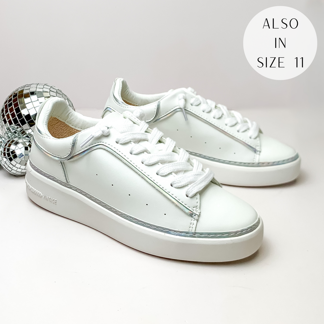 Pictured is a pair of white tennis shoes with white laces and a silver iridescent outline. These shoes are pictured on a white background with disco balls on the left side