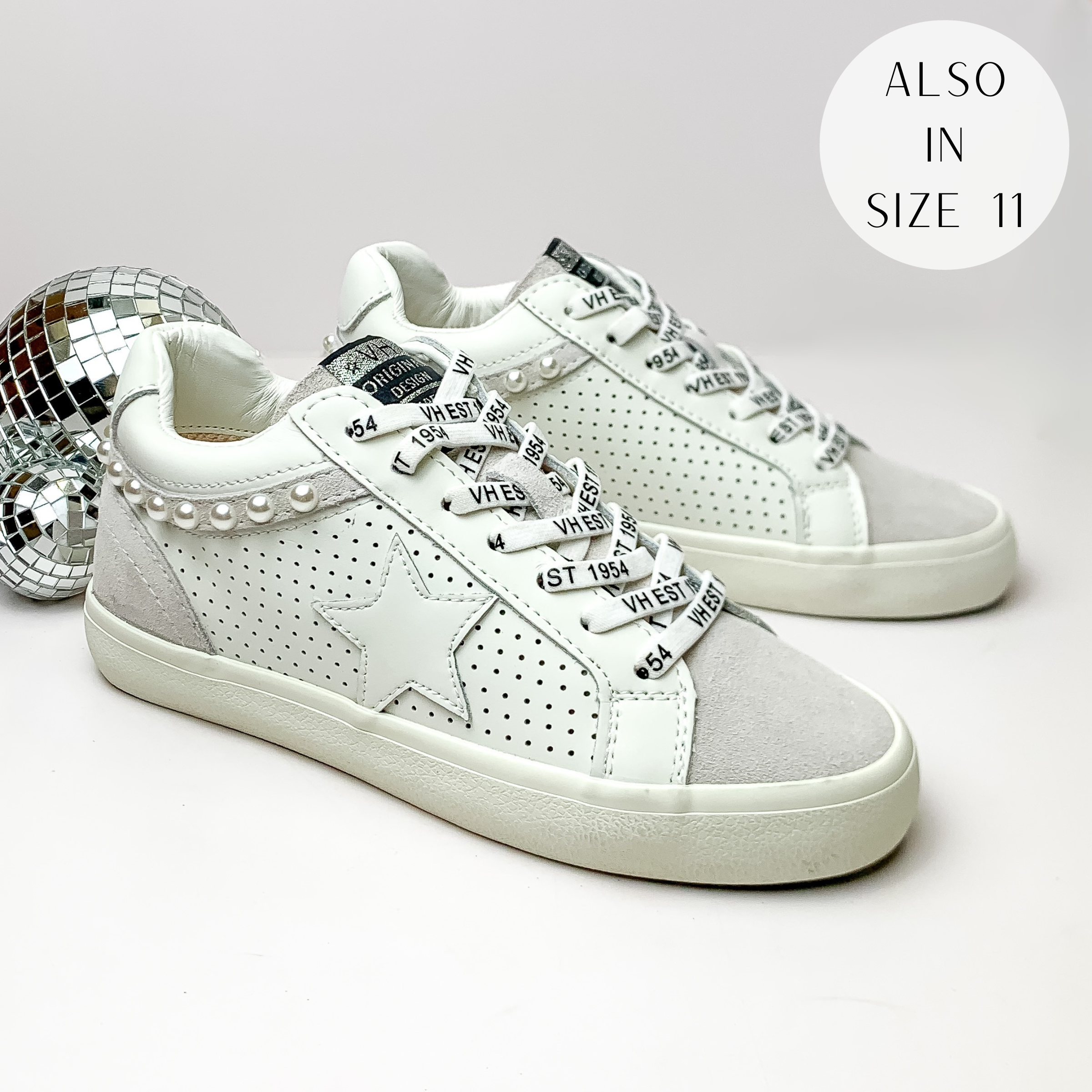 Pictured is a pair of white tennis shoes with white laces, grey detailing, and a faux pearl outline. These shoes also include a white star on the side. These shoes are pictured on a white background with disco balls on the left side