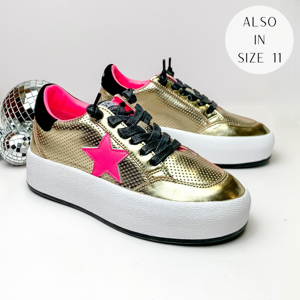 Pictured is a pair of gold tennis shoes with black laces, black detailing, and a hot pink star on the side. These shoes also include a white platform sole. These shoes are pictured on a white background with disco balls on the left side