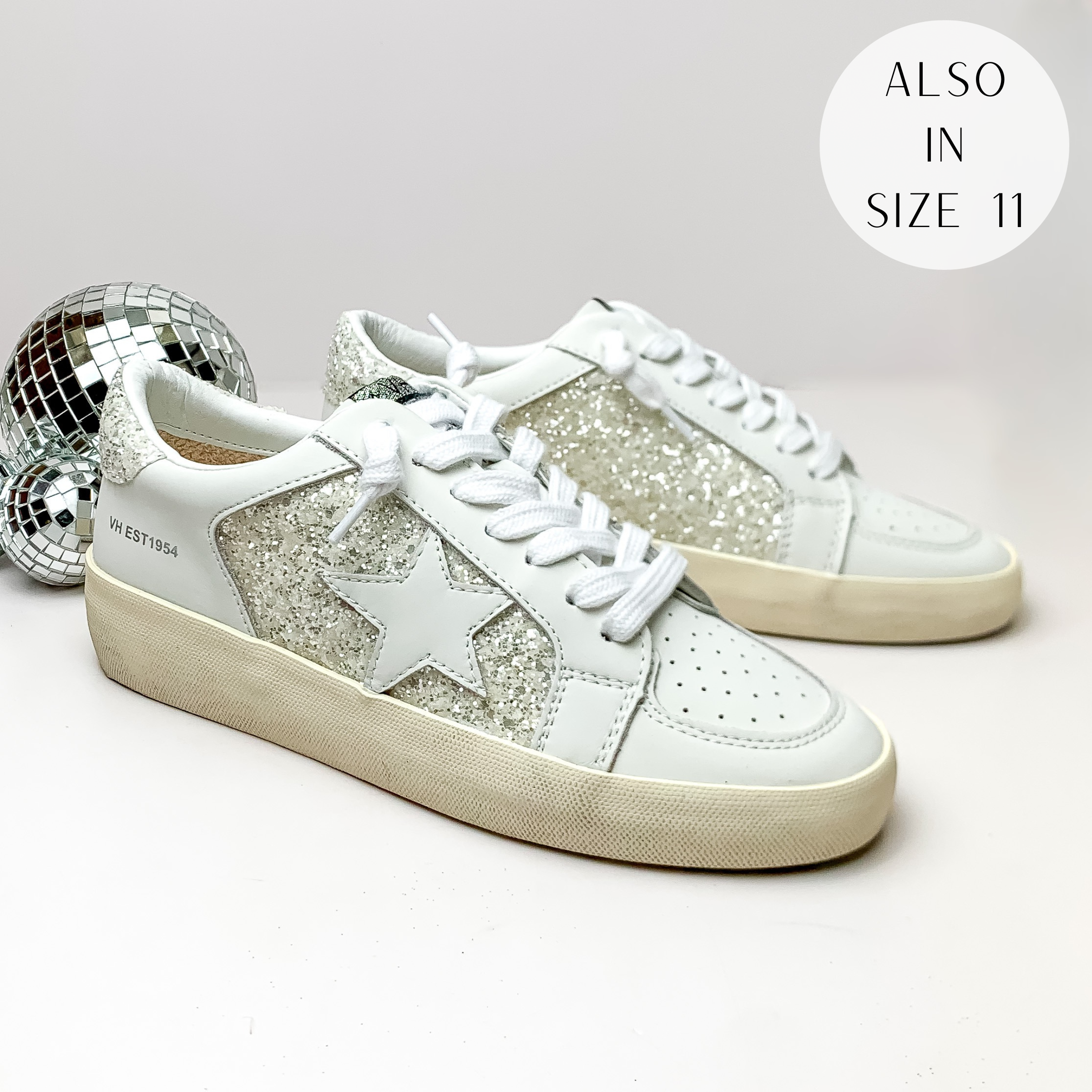 Pictured is a pair of white tennis shoes with a white star on the side and white glitter detailing. These shoes are pictured on a white background with disco balls on the left side.