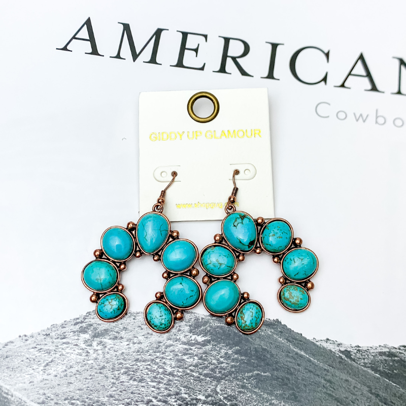 Squash Blossom Stone Earrings In Turquoise. Pictured on a white background with a western scene in the back.