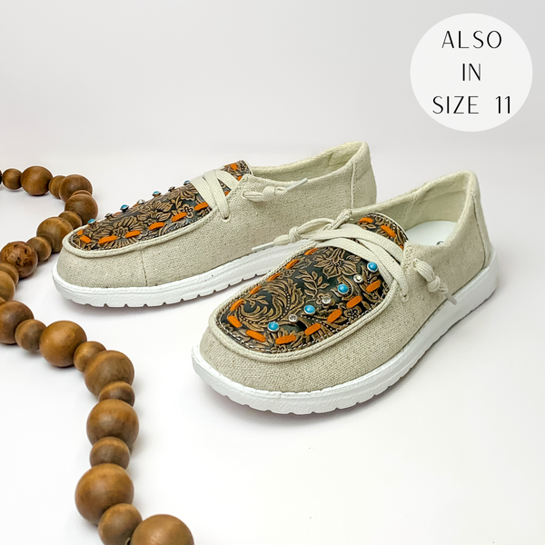 Beige canvas shoes with a leather tooled upper and beige laces. The leather upper has an orange stitched outline with blue and clear crystal studs on one side. These shoes are pictured on a white background with brown beads on the left side.
