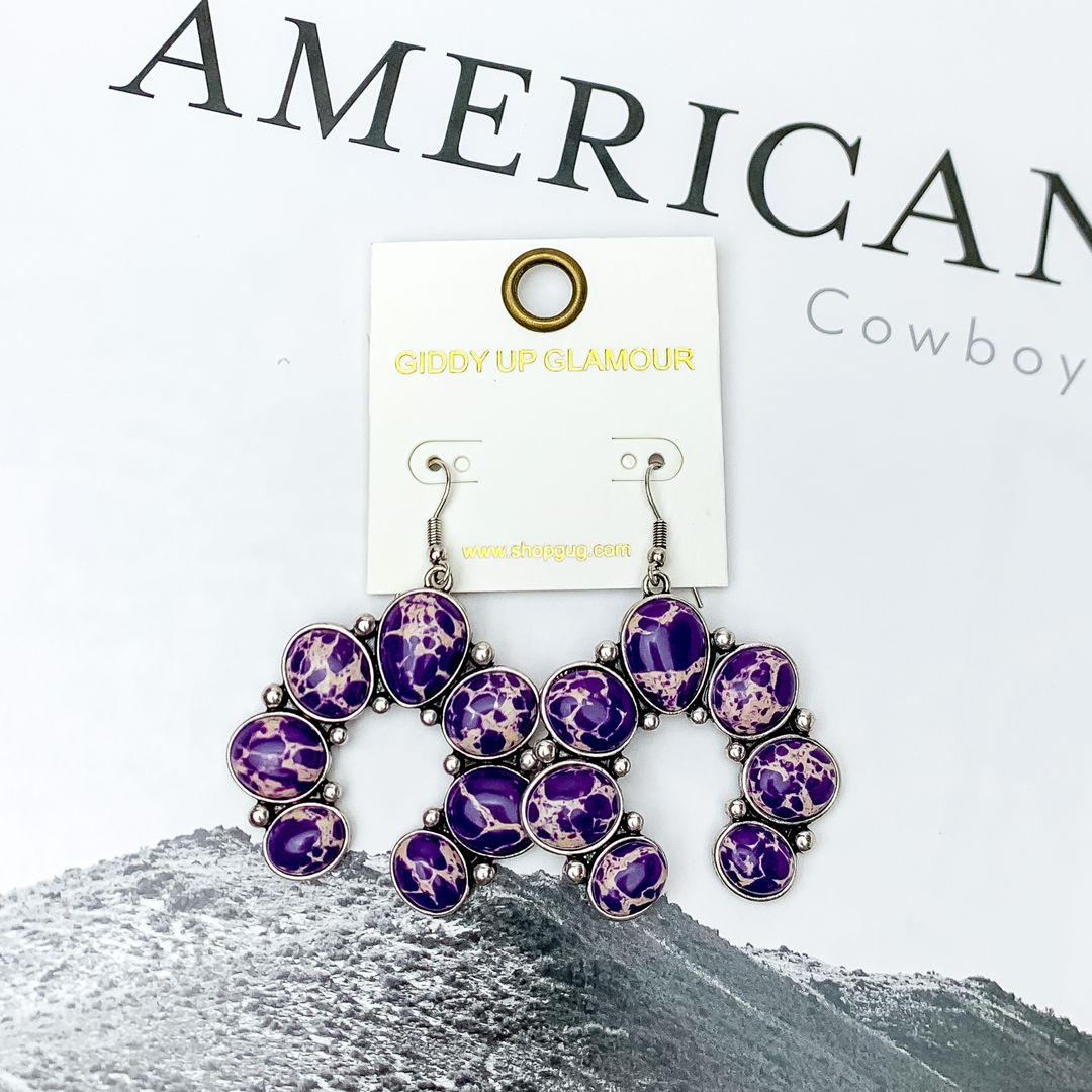 Squash Blossom Stone Earrings In Purple. Pictured on a white background with a western scene in the back.