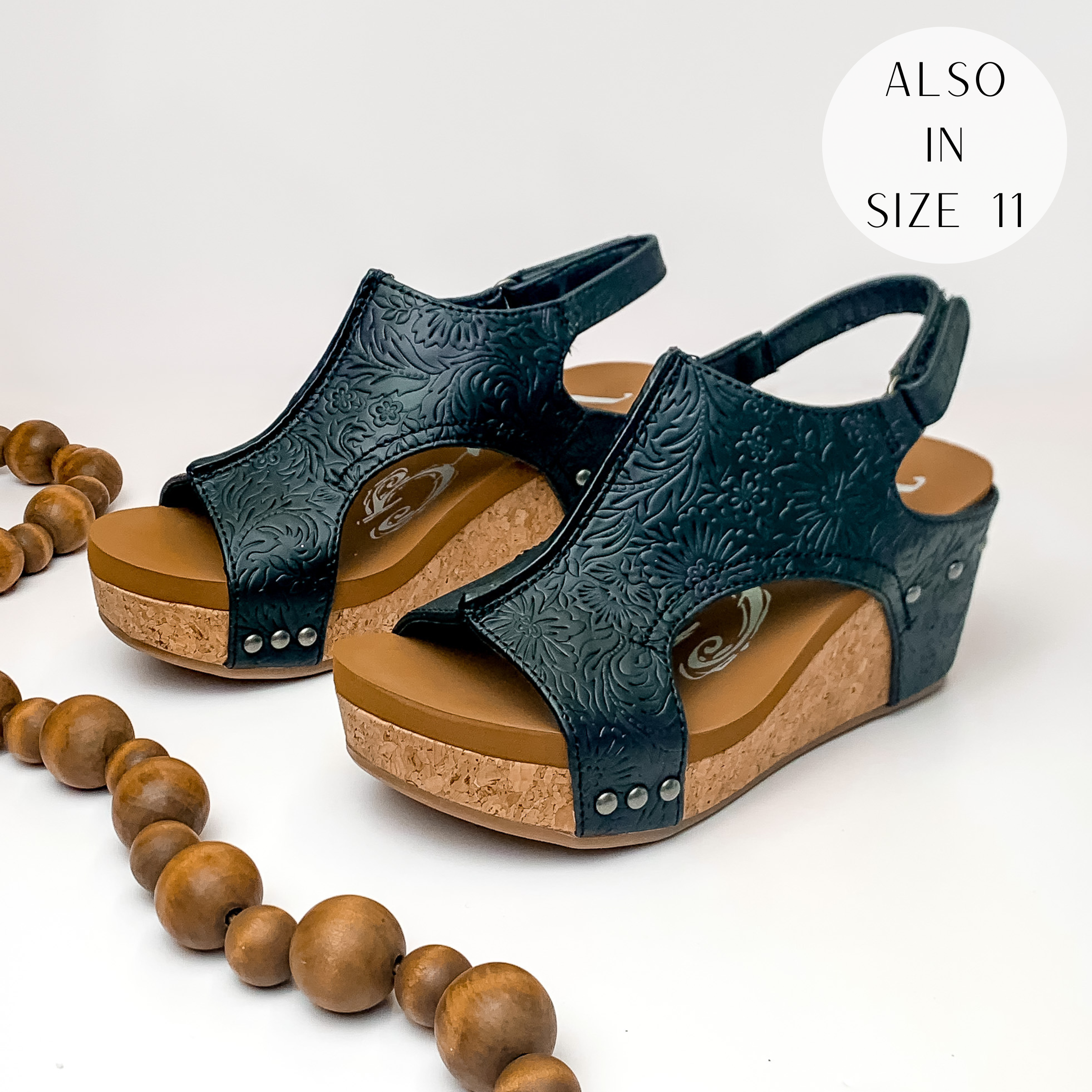 Black, leather tooled cork wedge sandals. These wedges also include charcoal silver studs and a velcro strap. These wedges are pictured on a white background with brown beads on the left side.