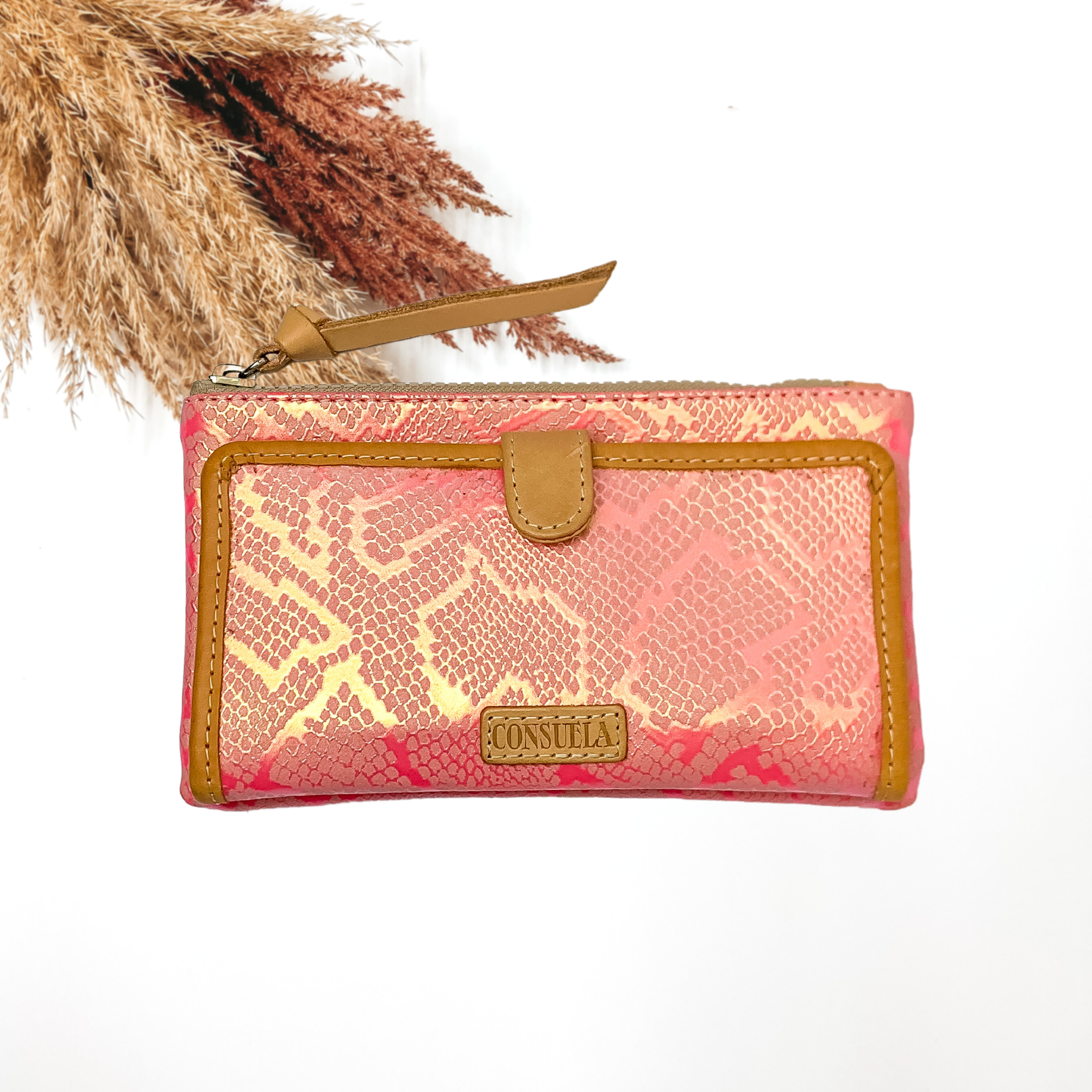 Pink, iridescent snakeskin print wallet with a front closure and top zipper. This wallet also includes a light tan tassel and a light tan outline on the front flap. This wallet is pictured on a whte background with brown pompous grass in the top left corner. 