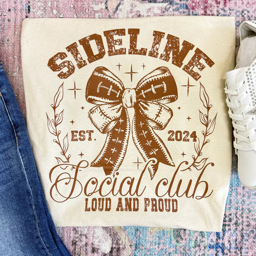 Beige graphic tee with 'Sideline Social Club' design featuring a football bow, text, and shoes on a textured surface.