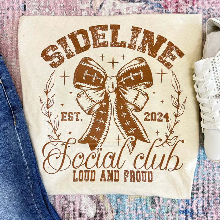 Beige graphic tee with 'Sideline Social Club' design featuring a football bow, text, and shoes on a textured surface.