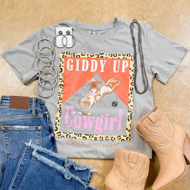Gray t-shirt with 'Giddy Up Cowgirl' design, jeans, boots, and accessories on a beige surface