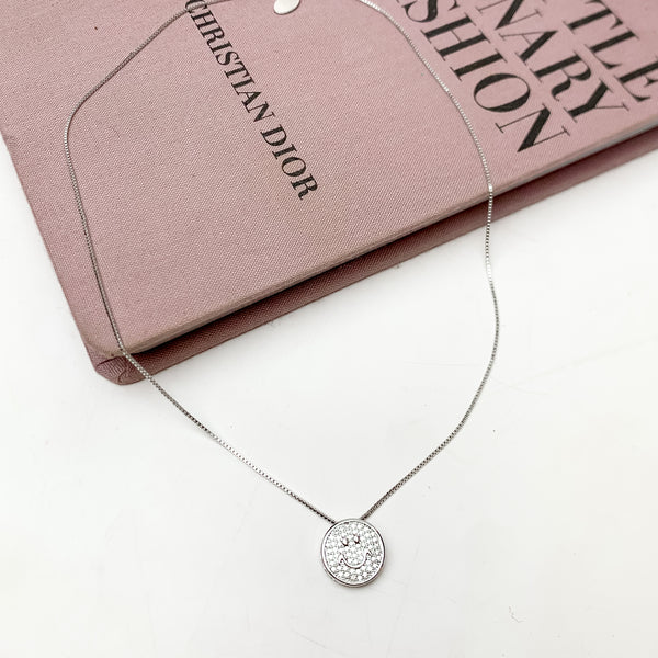 Silver Tone Chain Necklace With Clear Crystal Smiley Face. This necklace is pictured on a white background with part of it on a pink book.