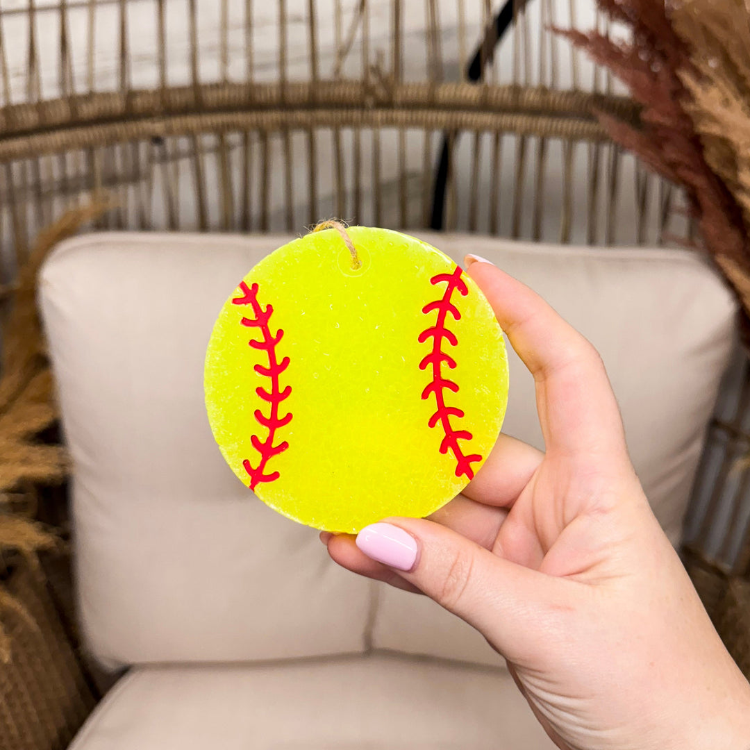 Background is a blurred wooden rustic chain. In the front and center is a hand holding a car freshie that is shaped and painted yellow to look like a softball including the red painted stitches. 