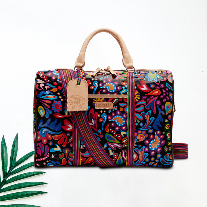 White background with palm leaf. Sophie bag with a vibrant Consuela Cloth exterior, natural leather handles, and a multicolored woven adjustable crossbody strap. Includes a leather bag tag, zipper closure, interior zipper and open pockets, and two exterior open pockets. Trimmed in natural leather.