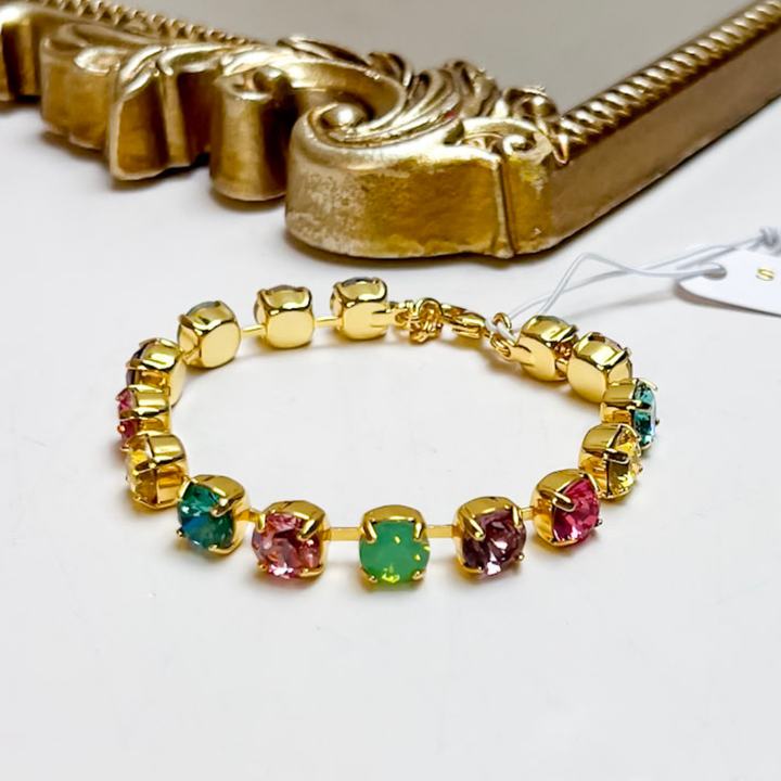 A gold-plated bracelet featuring multiple bezel-set gemstones in pink, green, and blue, displayed on a white surface with a decorative gold frame in the background.