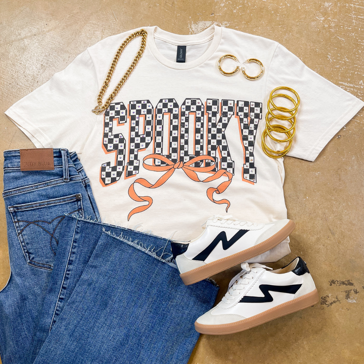 White t-shirt with 'Spooky' design, blue jeans, white sneakers, and gold accessories on a beige surface.