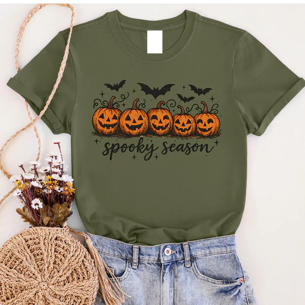 Green t-shirt with Halloween design featuring pumpkins and text, displayed with jeans and decorative items.