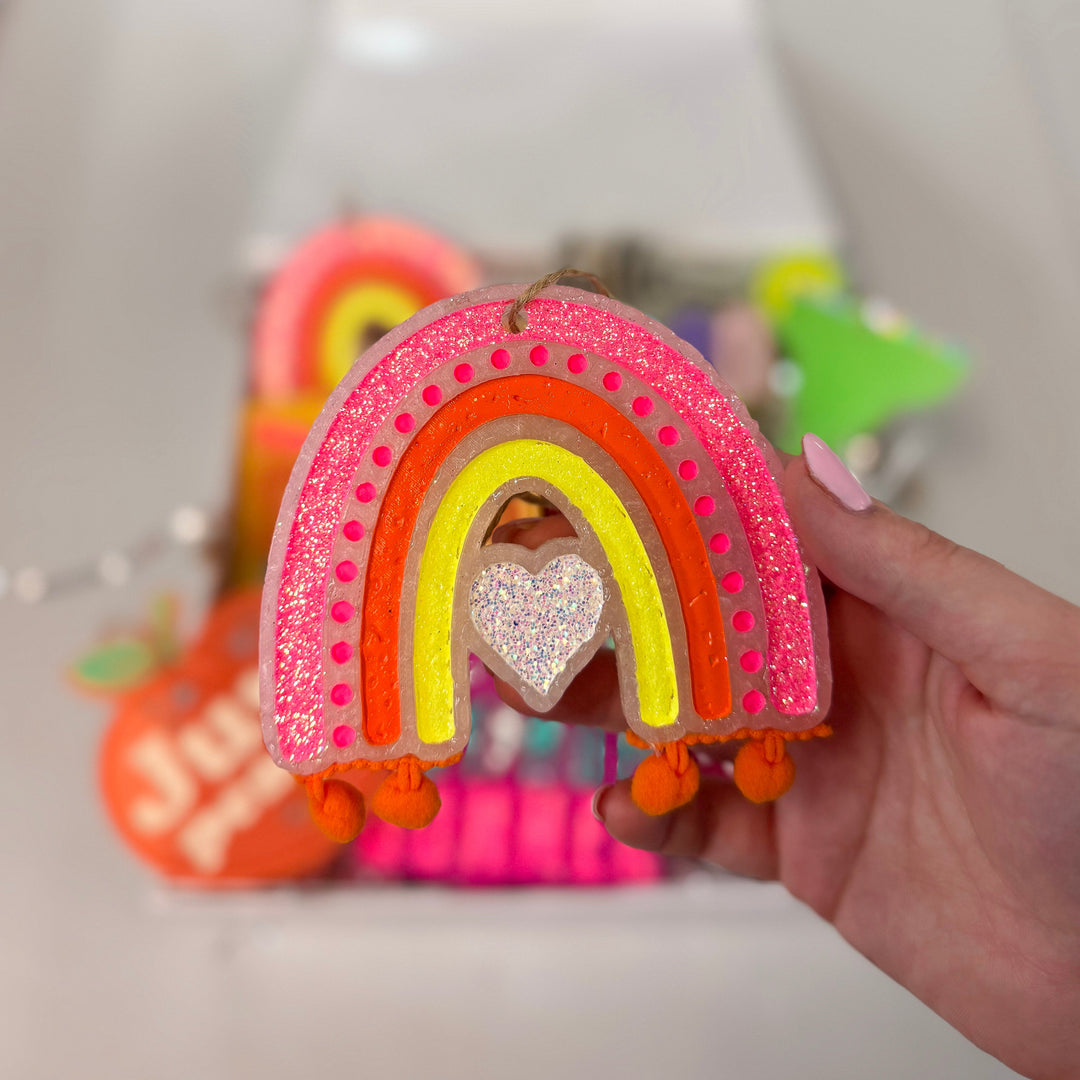 Rainbow car freshie. Each arch is a different color. In the center there is a silver sparkly heart. At both ends of the rainbow are orange pom poms. 