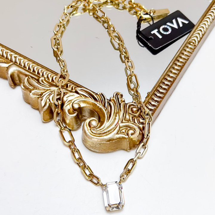 Gold necklace with intricate design and clear crystal pendant on a white background, featuring the brand &