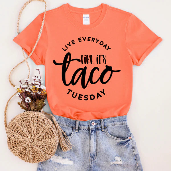 Coral t-shirt with 'Live Everyday Like It's Taco Tuesday' text, paired with denim shorts and a woven bag on a white background.
