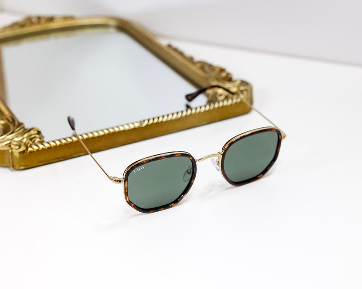 Sunglasses with gold frame and dark lenses on a white surface with a decorative gold frame in the background.