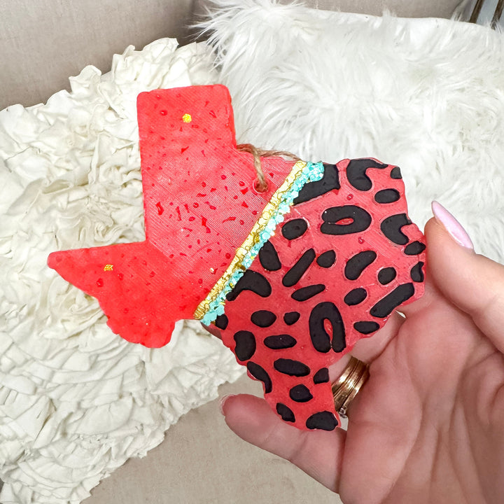 Hand holding a red and black patterned car freshie against a white textured surface.