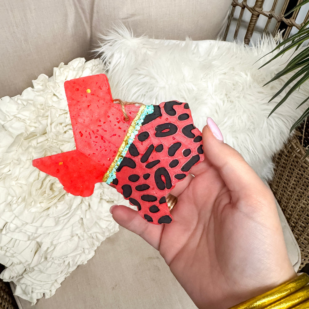 Hand holding a car freshie with a red and black pattern, sitting on a textured surface.