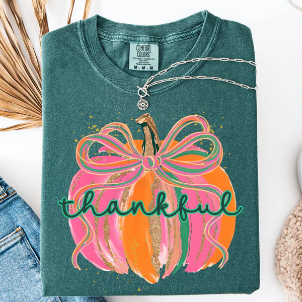 Teal shirt with a colorful pumpkin design and 'thankful' text, displayed with denim jeans and dried leaves.