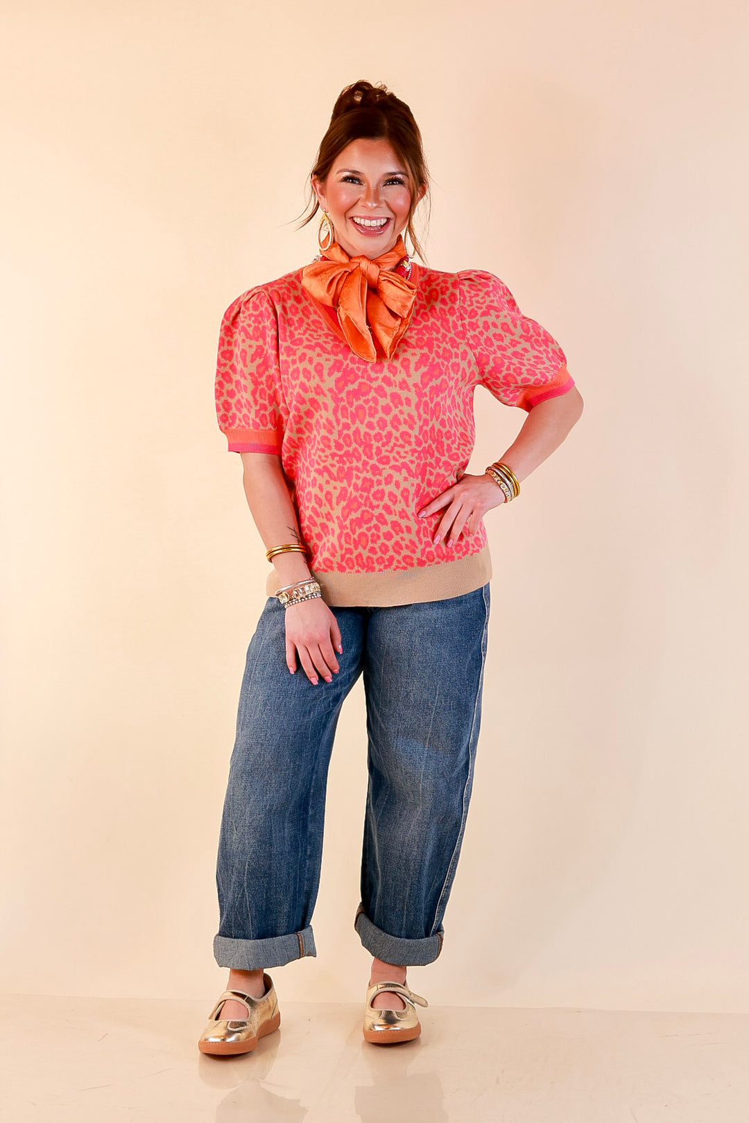 Model wearing a pink and orange leopard print puff sleeve sweater on a tan base with an orange silk scarf, cuffed wide-leg jeans and gold flats.