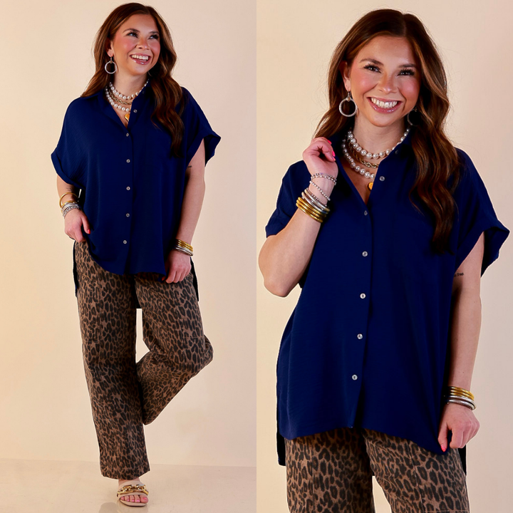 Model is wearing Woven Airflow Button Front Top with Rolled Cuff Short Sleeves and Front Pocket in Navy Blue with Jeans against a beige backdrop.