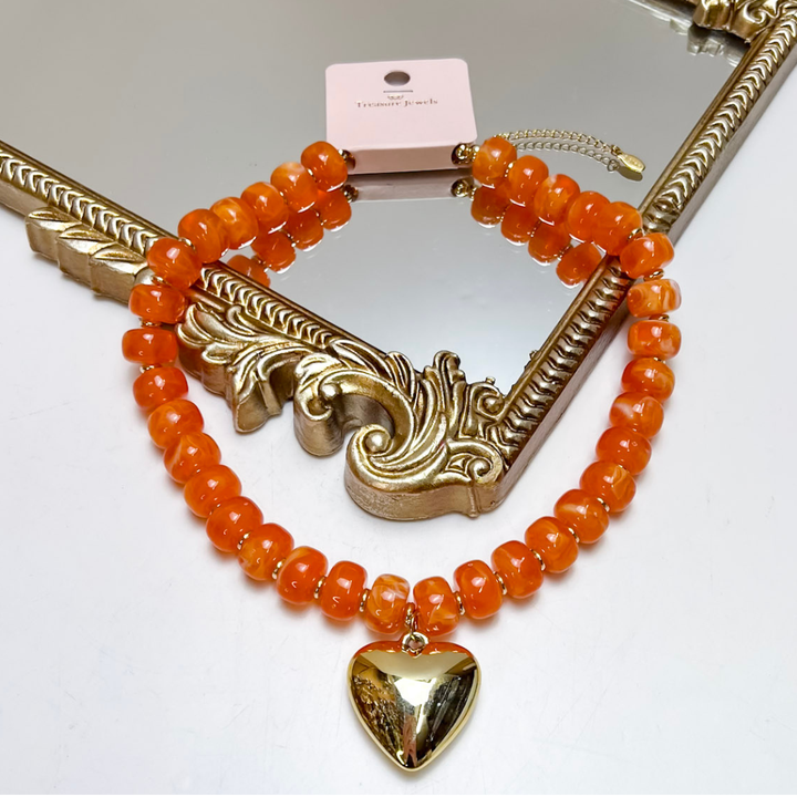An orange beaded necklace with a gold heart pendant, displayed on an ornate gold mirror frame with a pink tag attached.