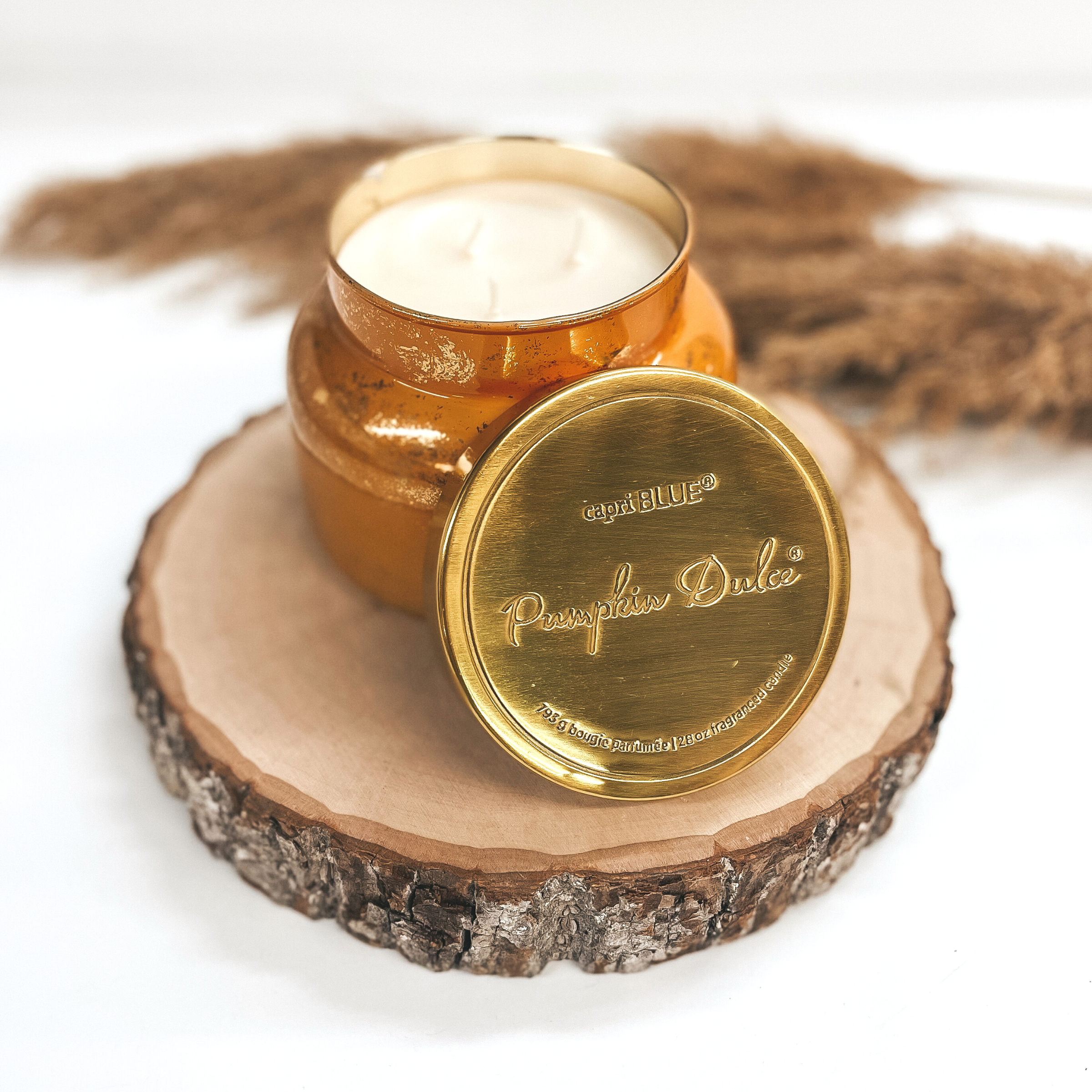 Orange candle with gold flecks and a gold lid pictured on a white background on a piece of wood with brown pompous grass in behind the candle. 